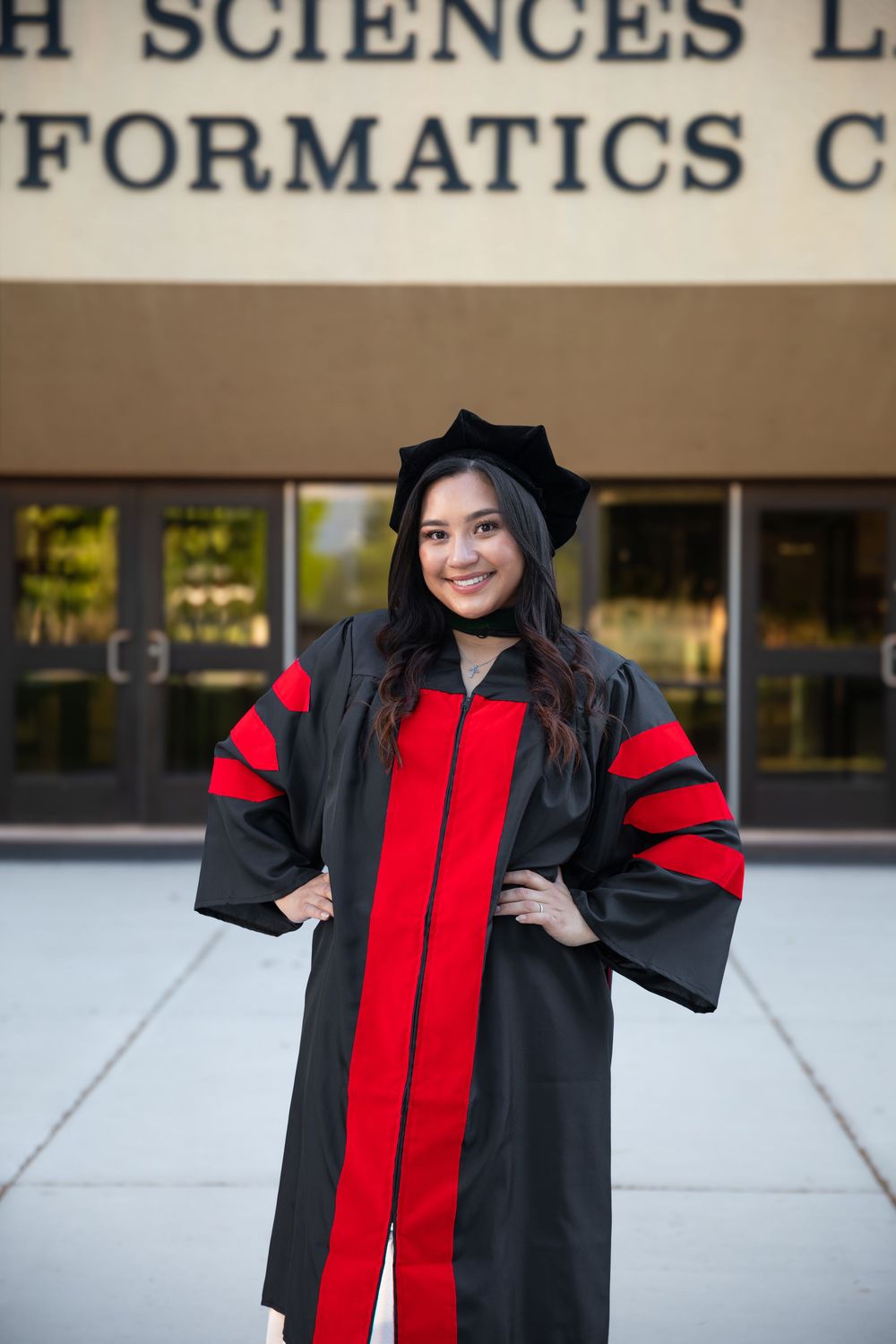 A smiling woman in black and red doctoral graduation gown and cap poses confidently in front of a university building.