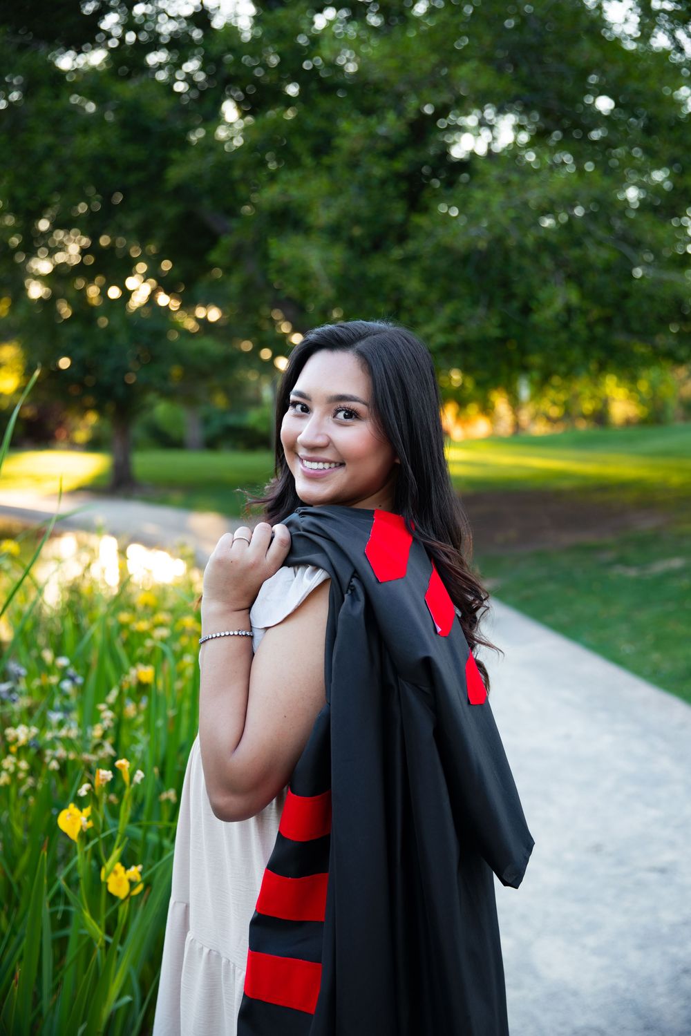 Smiling young woman in graduation gown over red striped dress, posing outdoors in a lush green park setting.