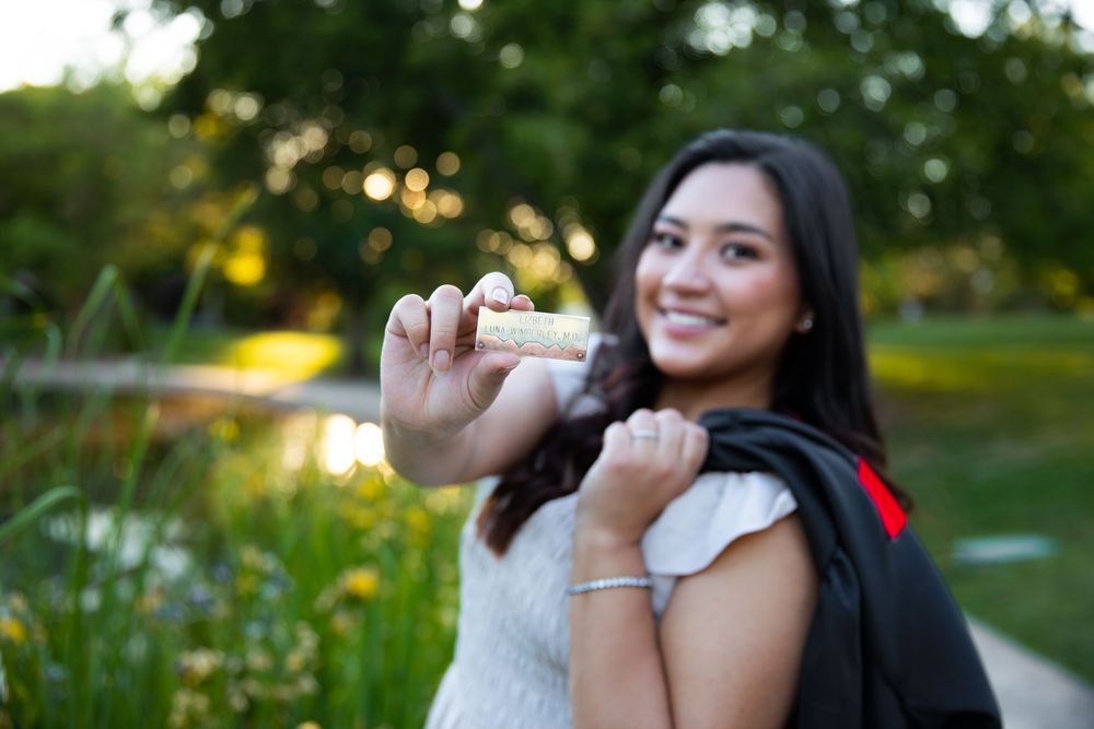 Smiling female graduate holding diploma in park wearing cap and gown, celebrating academic achievement outdoors.