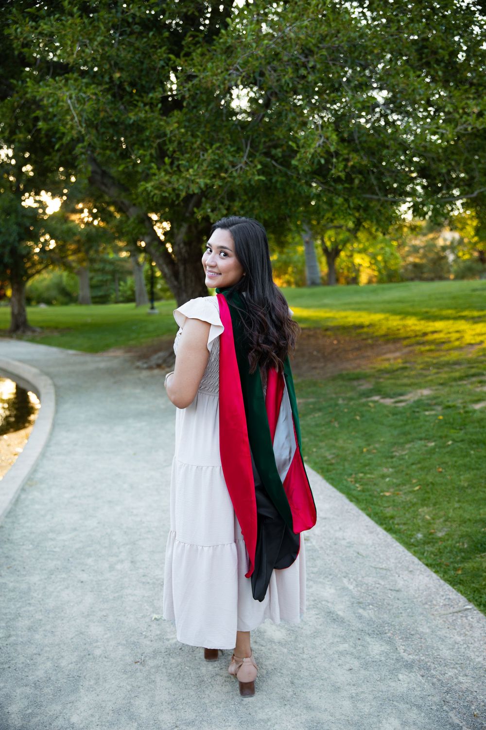 Young woman in white dress with red and green graduation stole walks along park path, glancing back with a smile.