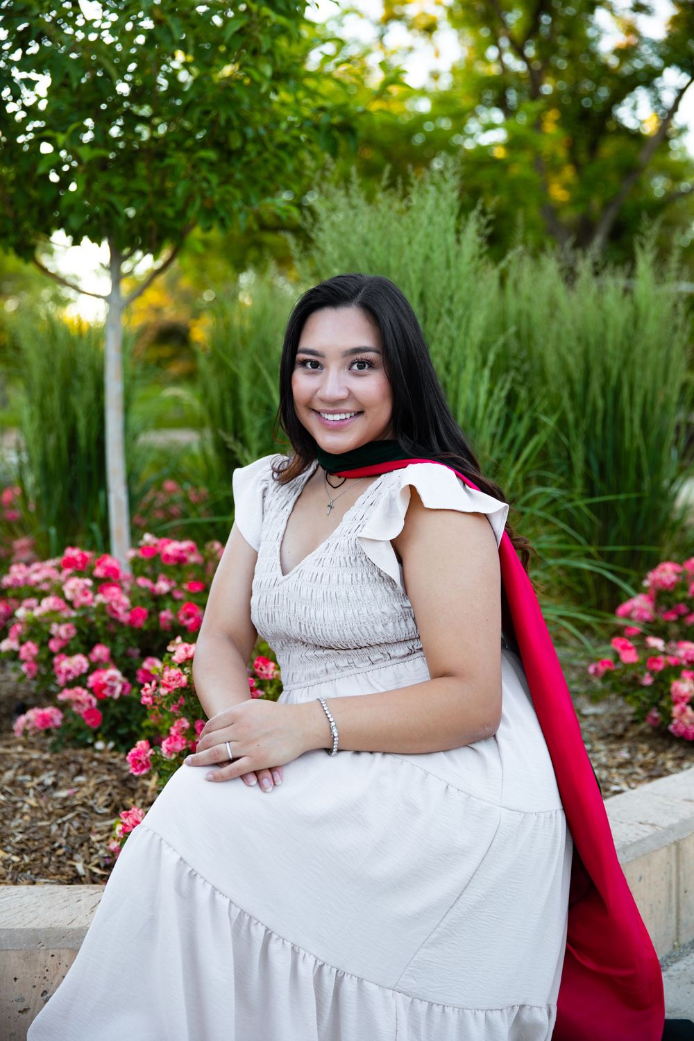 Young woman in white dress and red cape smiles while seated in a garden with pink roses and green trees.