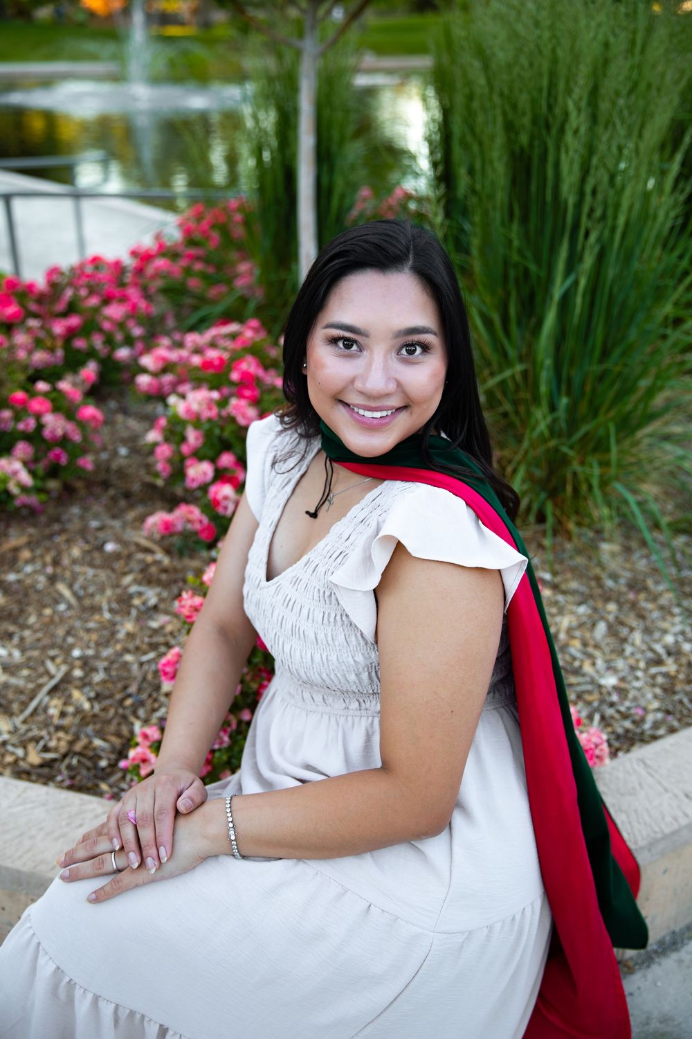 Smiling young woman in white dress with graduation stole sits outdoors near pink flowers in a garden setting.