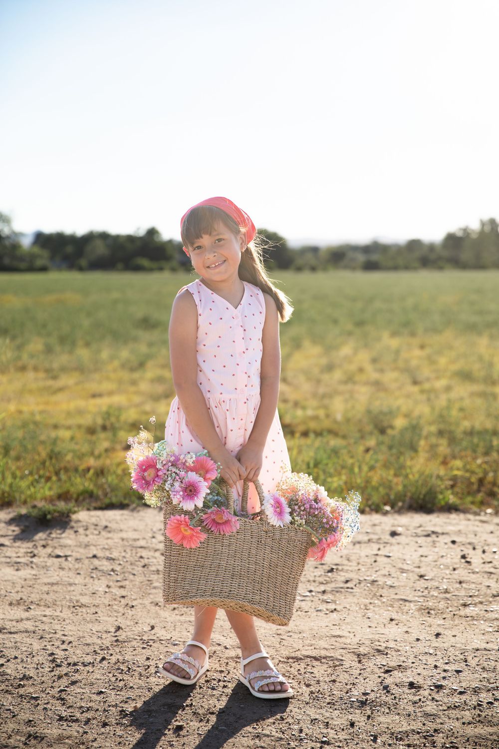 Young girl in white dress holding a wicker basket of pink flowers in a sunny field.