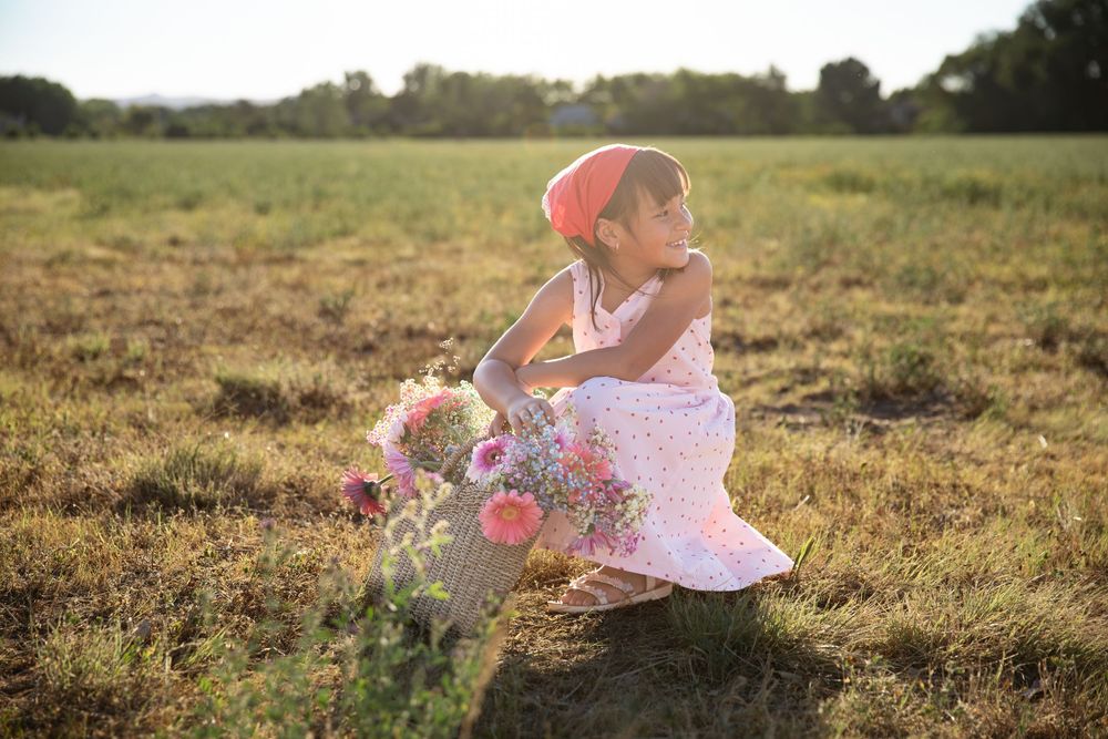 Young girl in floral dress and red hat crouching in sunny field holding bouquet of pink wildflowers.