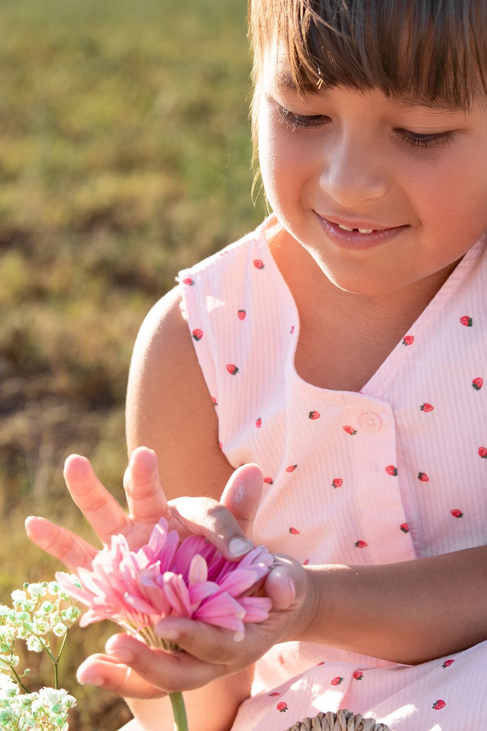 Young girl in pink ladybug dress smiling while gently holding a pink flower outdoors in sunlight.