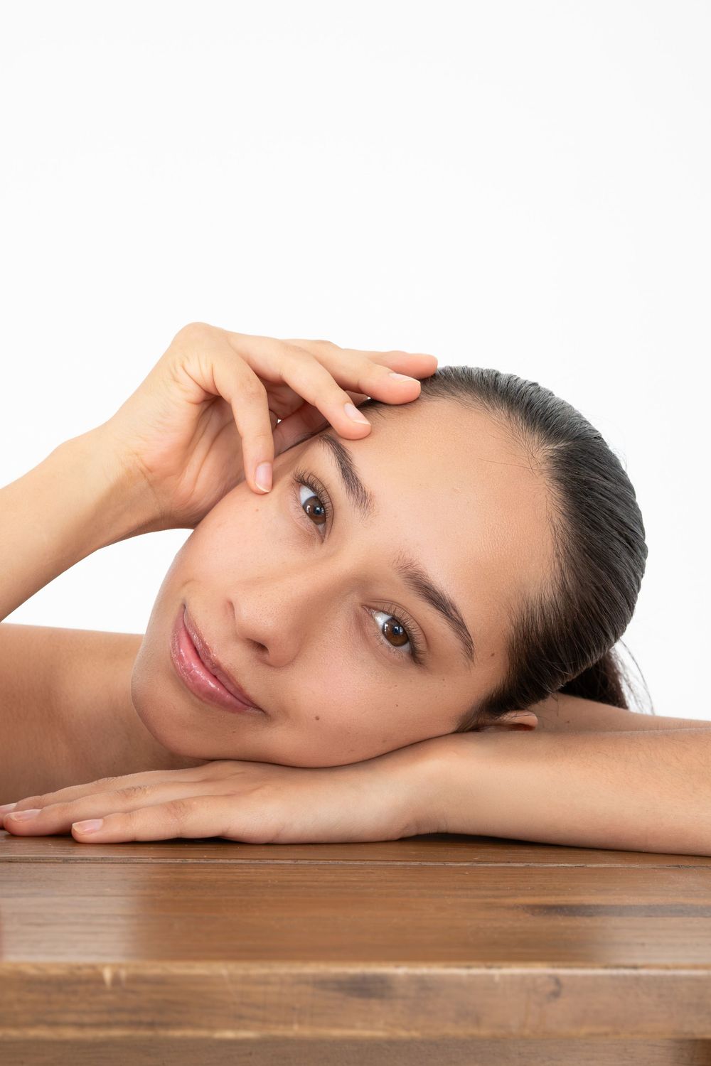 Young woman with natural makeup rests her chin on folded hands atop a wooden table, gazing softly at the camera.