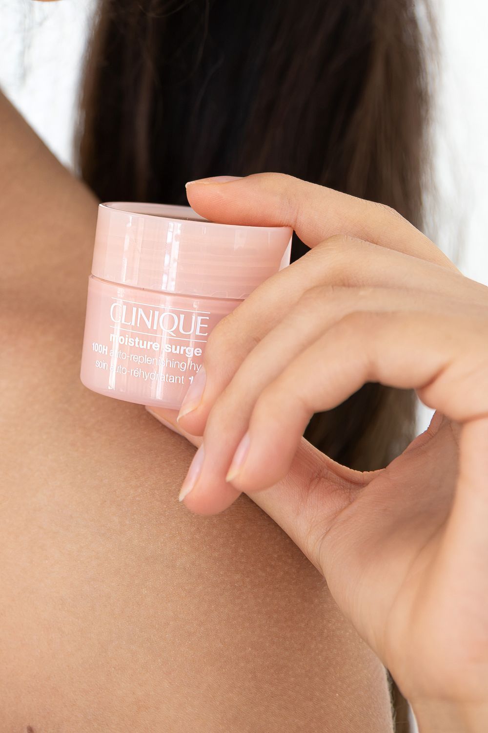 Hand holding a pink Clinique Moisture Surge moisturizer jar close to a woman's bare shoulder.