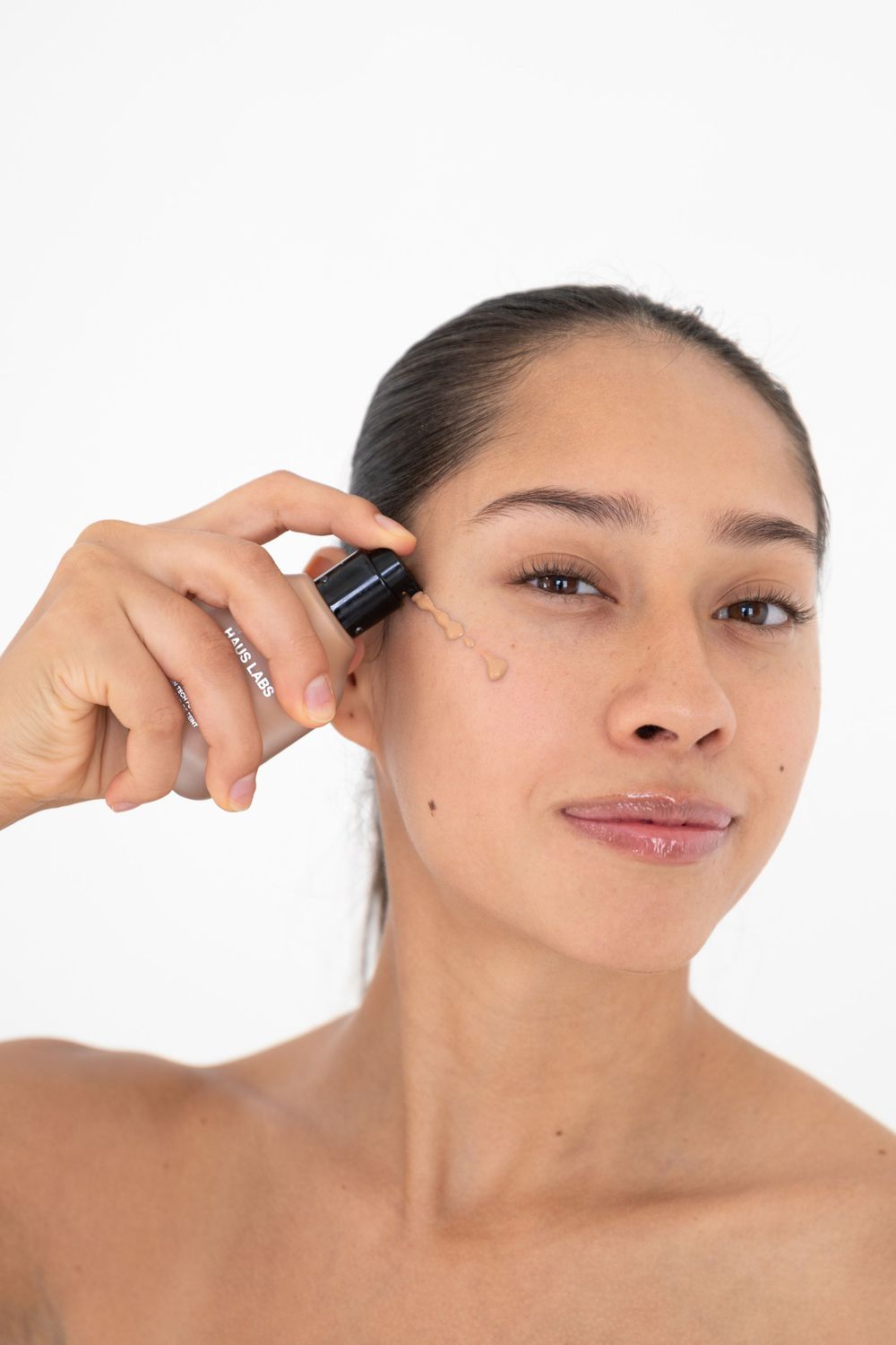Woman applying facial serum with dropper to cheek, showcasing skincare routine on white background.