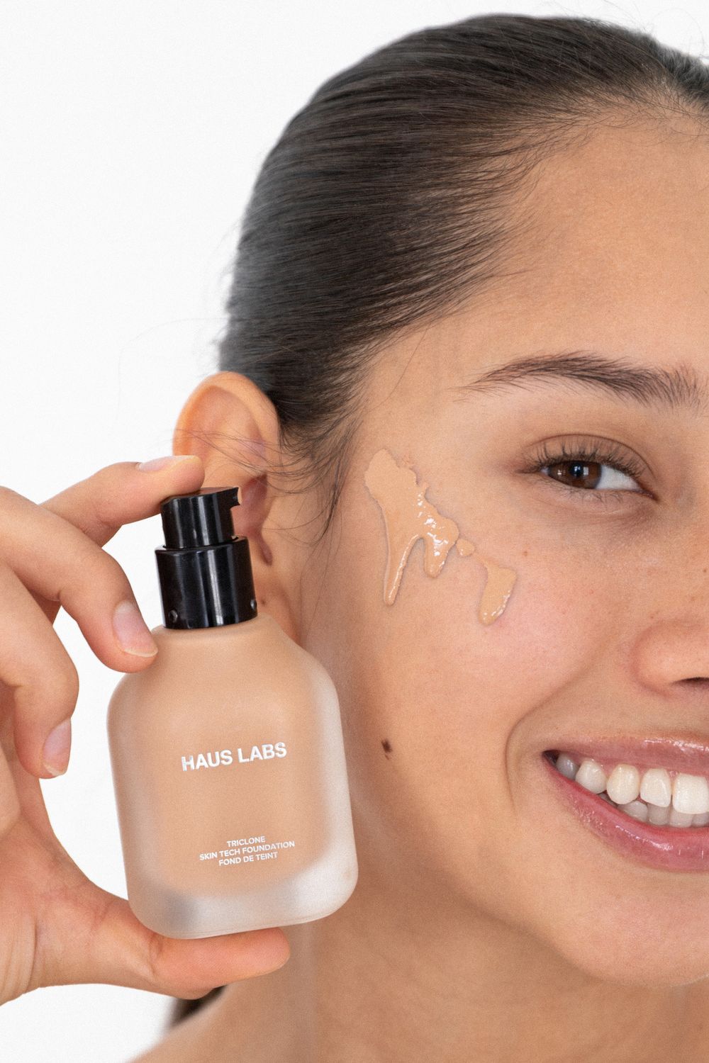 Woman smiling while holding Haus Labs foundation bottle with product swatches applied to her cheek.