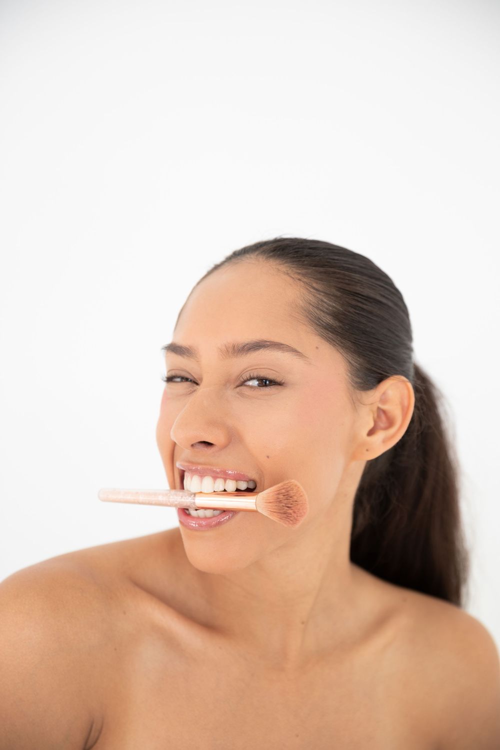 Smiling woman with dark hair holding a makeup brush in her mouth against a white background.