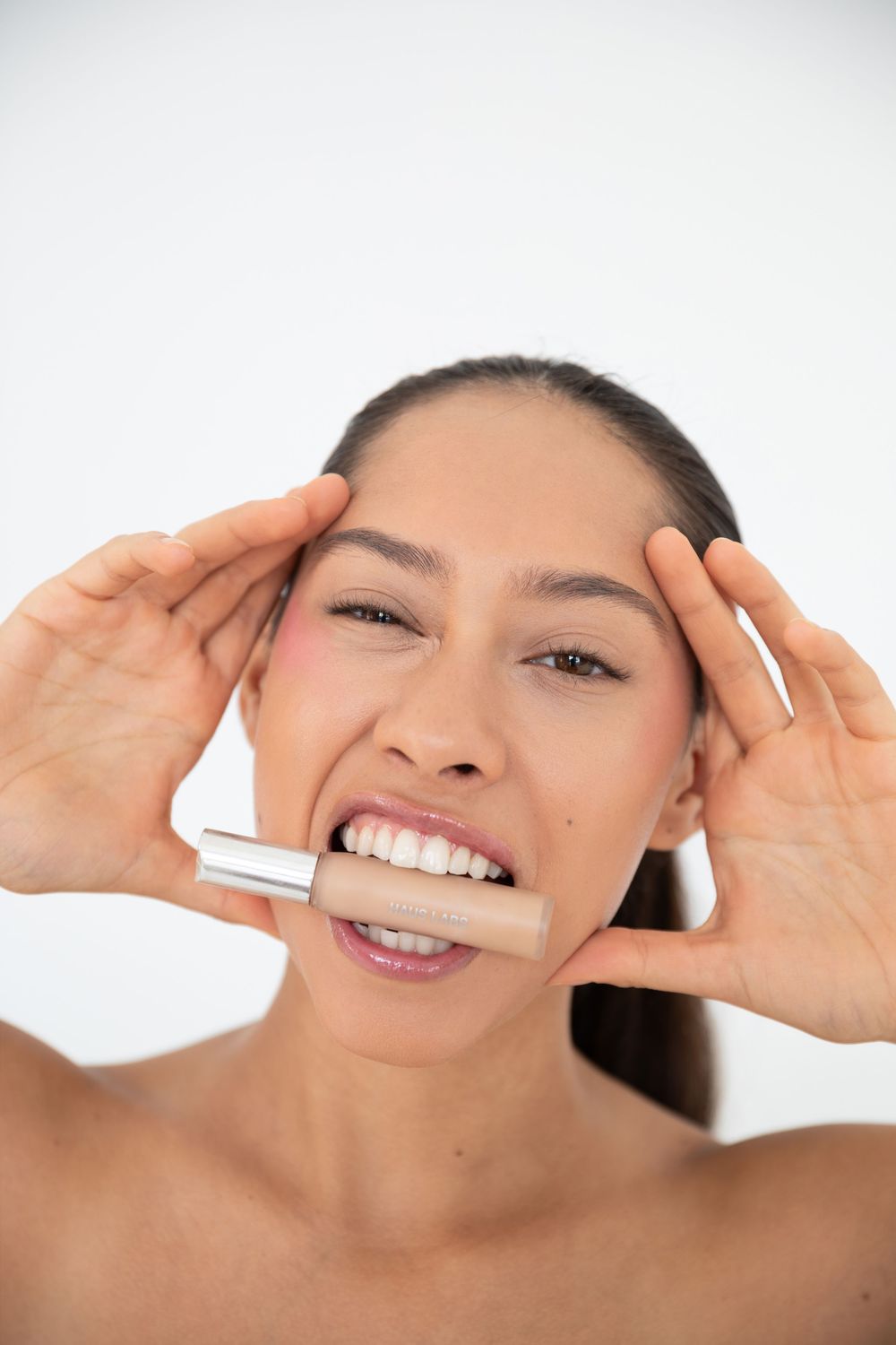 Woman holding a makeup foundation bottle in her teeth with hands framing her face against white background.