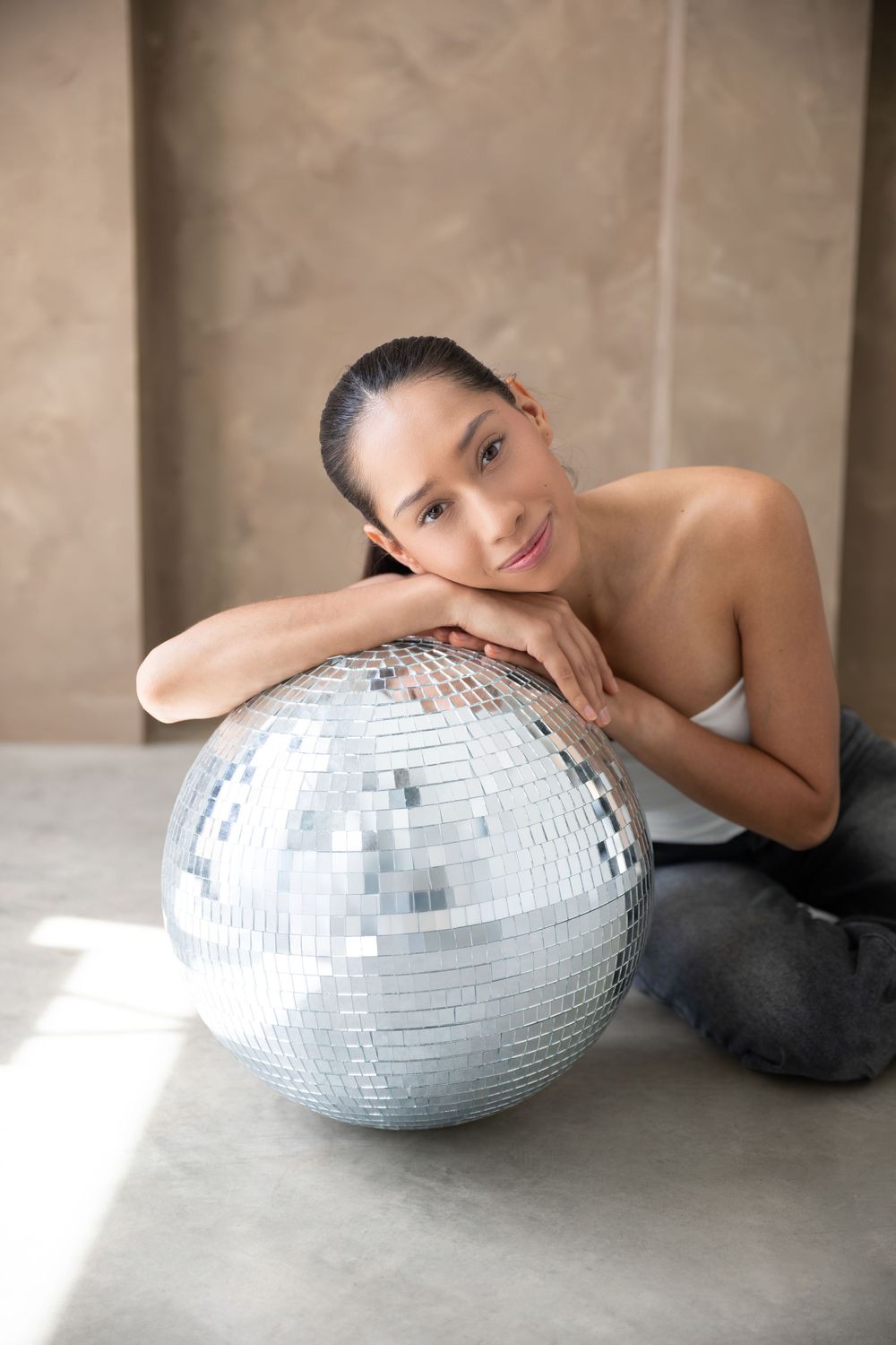 Young woman resting her chin on a large silver disco ball while sitting on the floor, smiling at camera.