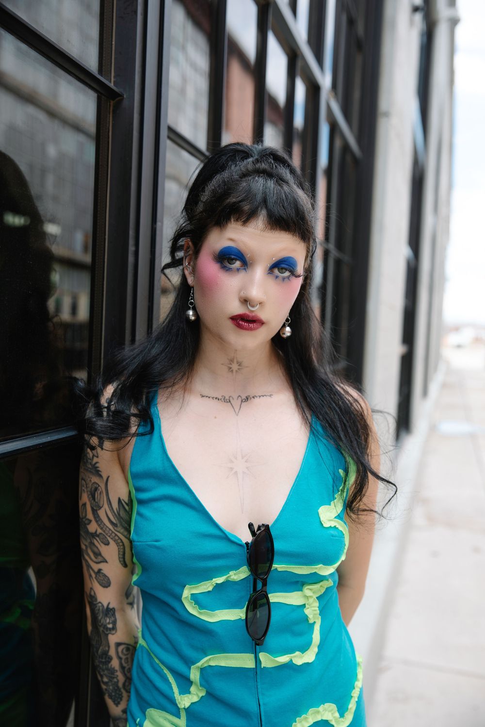 Woman with colorful eye makeup, tattoos, and teal dress leans against a dark building exterior.