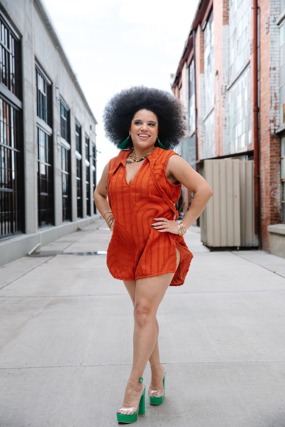 Woman in orange mini dress and green heels with afro hairstyle posing confidently in urban alleyway.