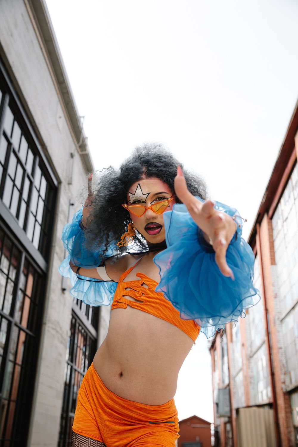 Woman in orange crop top and skirt with blue ruffles and star sunglasses poses boldly in urban alleyway.