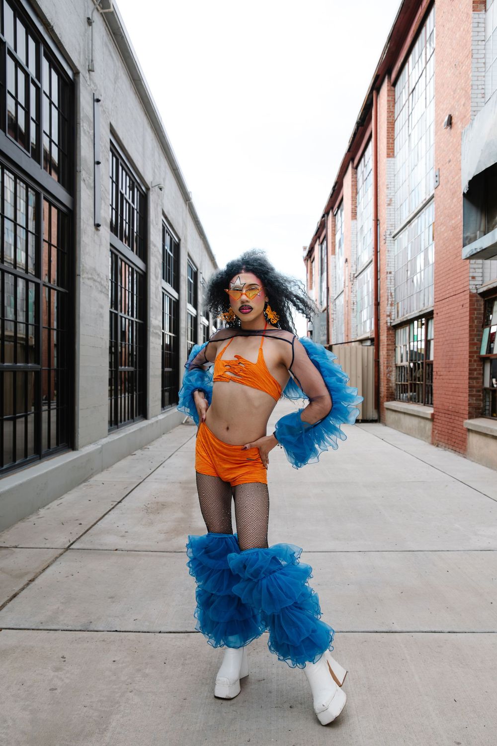 Person in orange crop top and shorts with blue tulle accessories and white boots posing in urban alley.