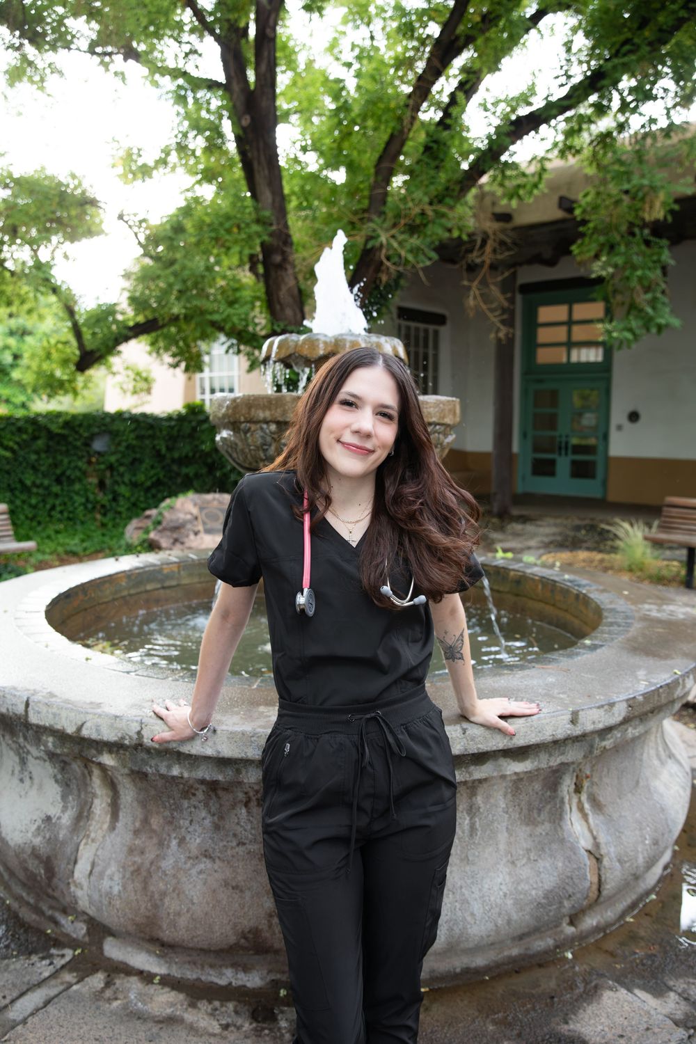 Young woman in black scrubs with stethoscope smiling beside a stone fountain in a lush garden courtyard.