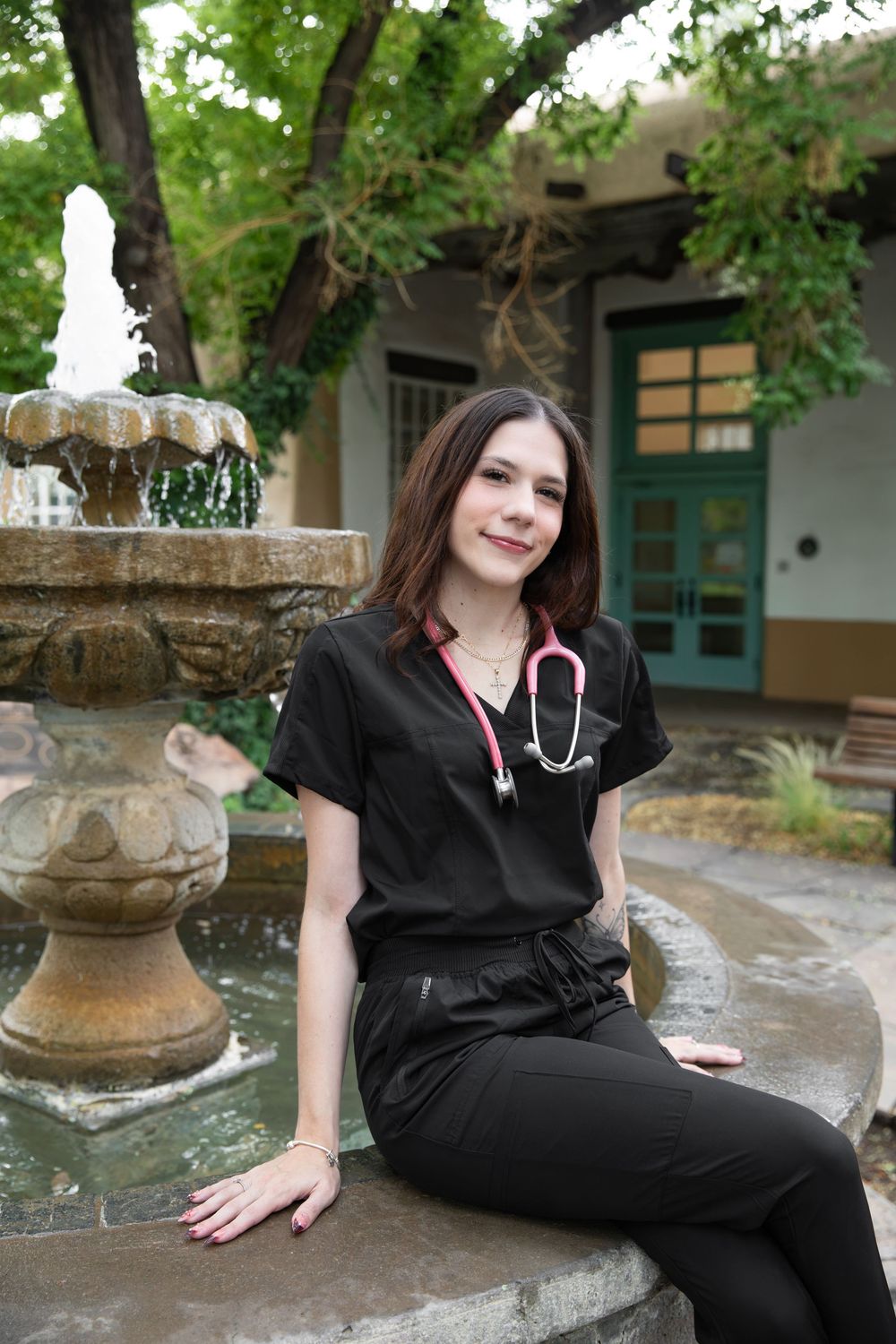Young female nurse in black scrubs with pink stethoscope sitting by stone fountain outdoors.