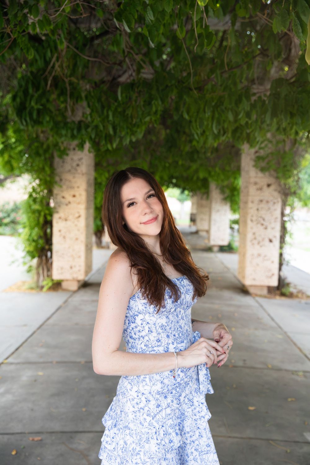 Young woman in blue floral sundress smiling under vine-covered pergola columns in outdoor garden setting.