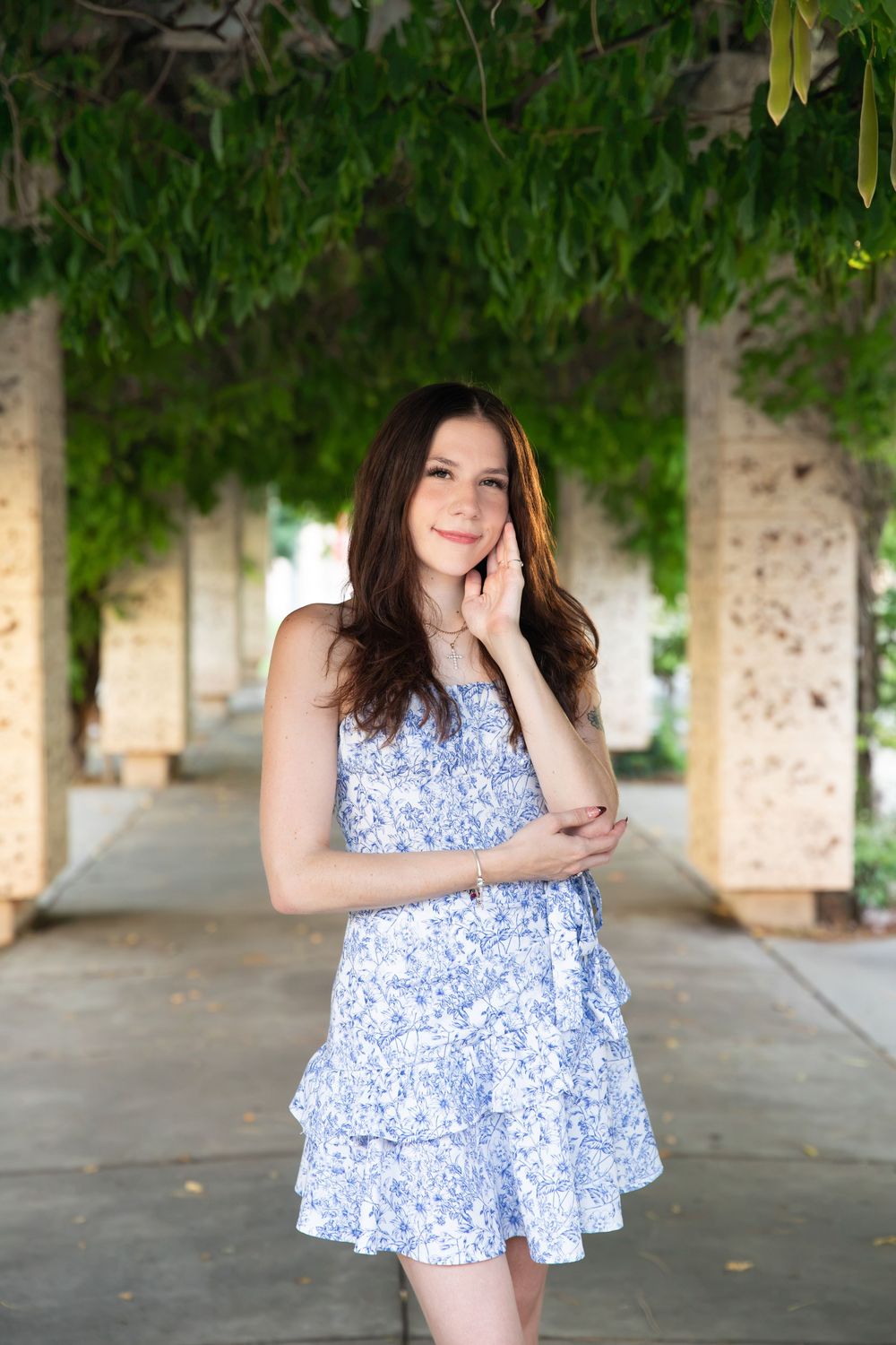 Young brunette woman in blue floral sundress posing under green vine-covered pergola columns.