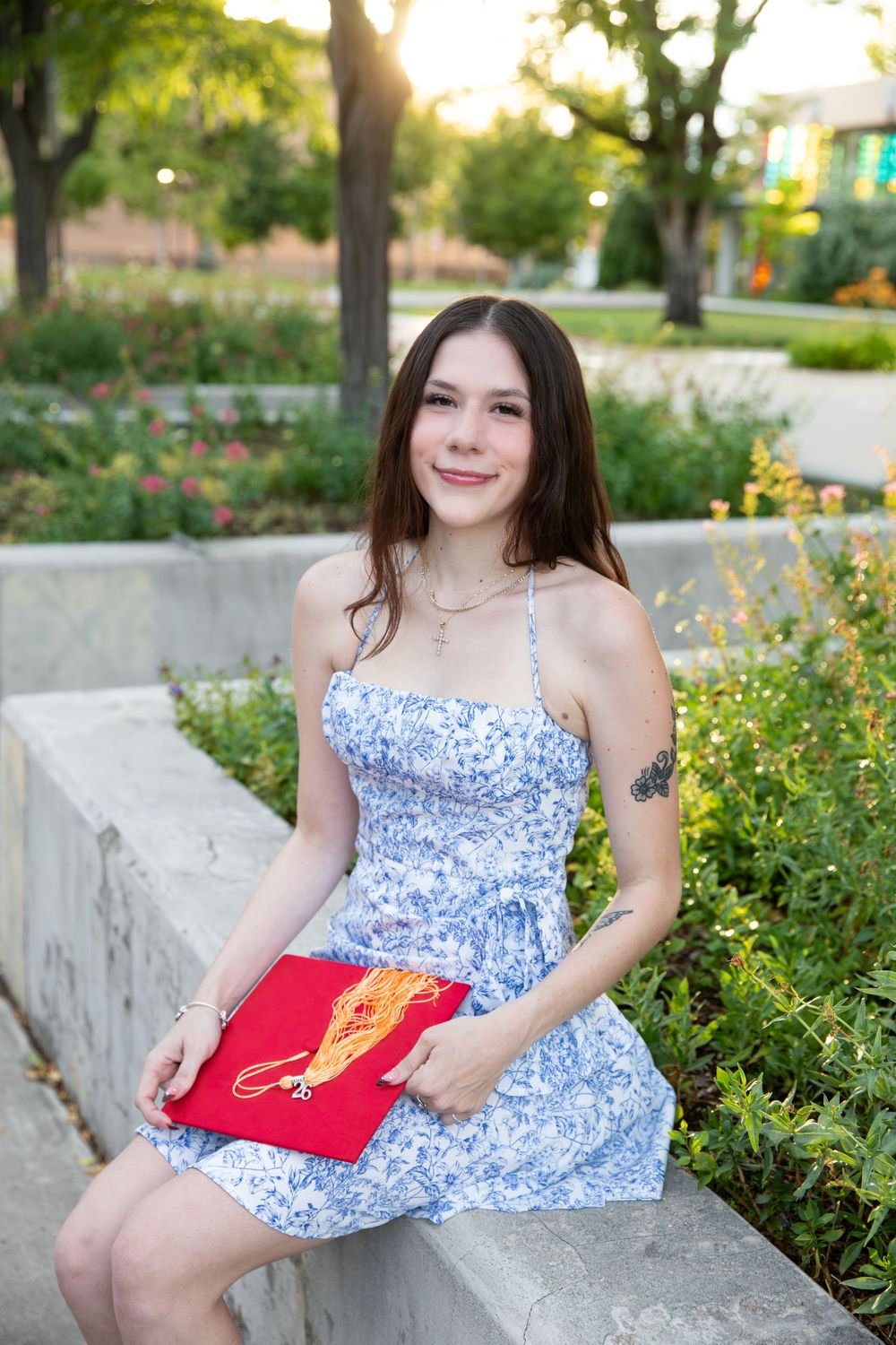 Smiling young woman in floral dress holding red graduation cap seated on stone wall outdoors.