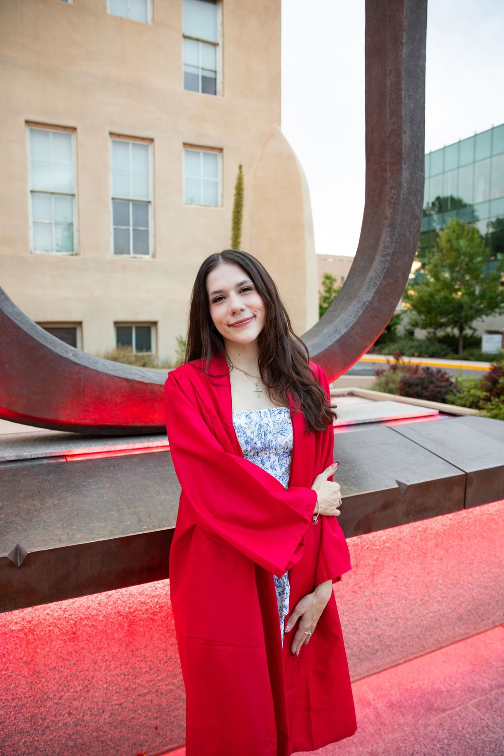 Young woman in red graduation gown posing in front of a large metal sculpture on a university campus.