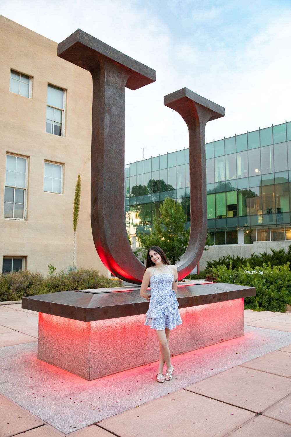 Young woman in floral dress posing by illuminated red U sculpture on university campus at dusk.