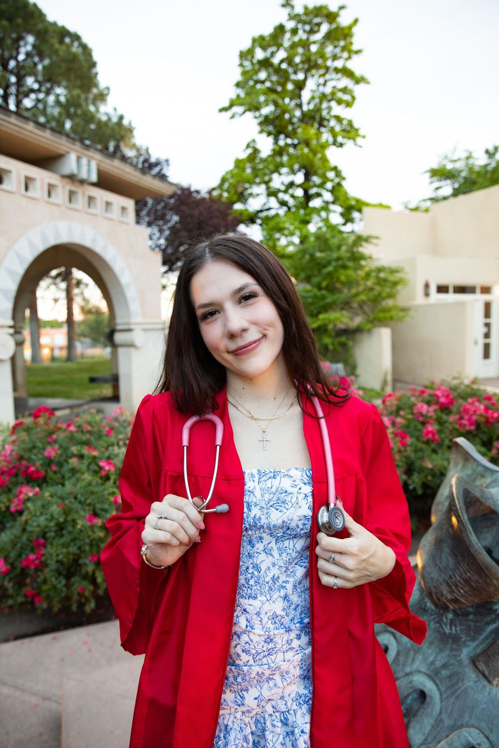 Young woman in red graduation gown holding a stethoscope, smiling outdoors near flowering gardens.