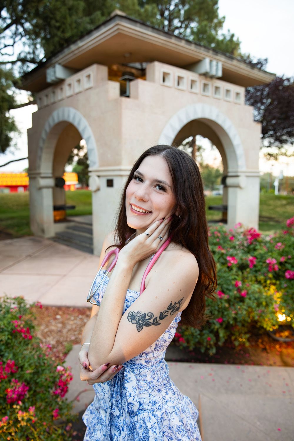 Smiling young woman with tattoos in blue floral dress poses in front of stone archway gazebo surrounded by pink flowers.