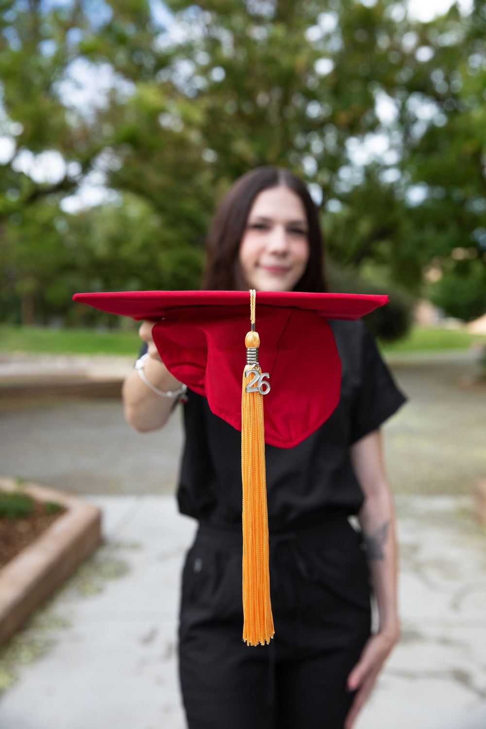 Young woman in black gown holding red graduation cap with gold tassel toward camera outdoors.