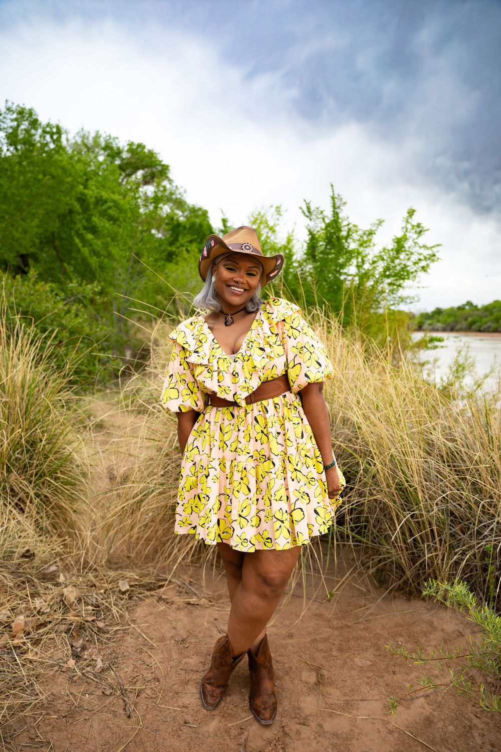 Woman in yellow floral dress and cowboy boots poses outdoors near river with green trees and cloudy sky.