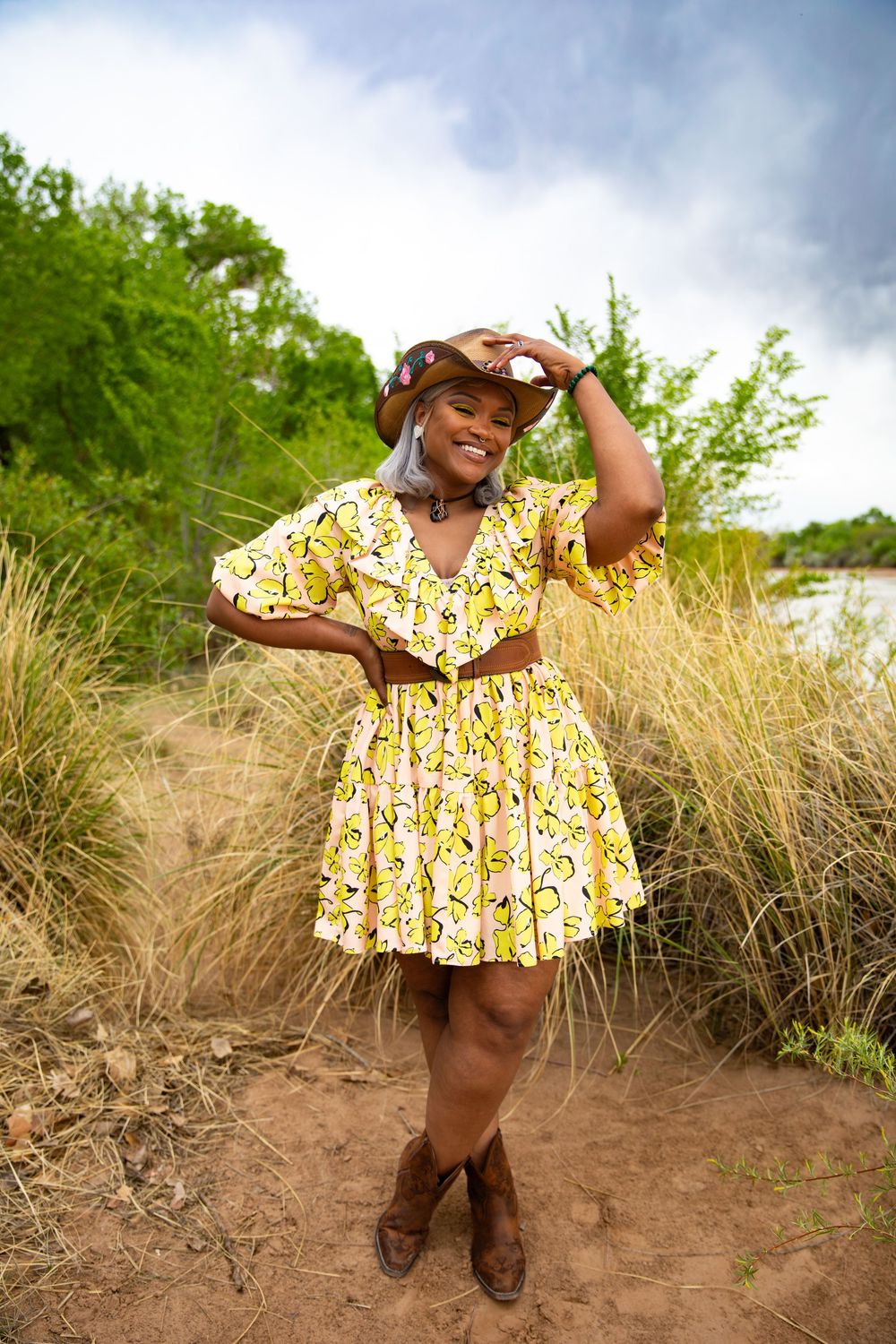 Smiling woman in yellow floral dress, cowboy hat, and boots poses outdoors among tall grass with trees in background.