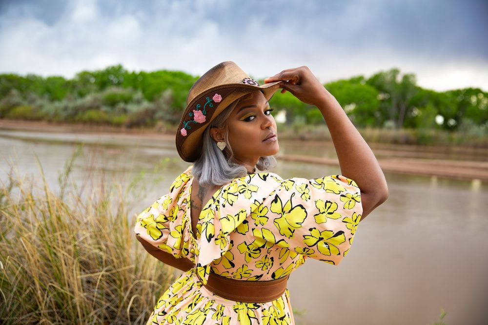 Woman in yellow floral dress and tan hat poses near a lake with lush greenery in the background.