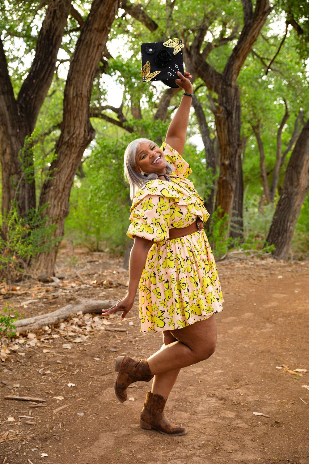 Woman in yellow floral dress and cowboy boots joyfully raises a black butterfly hat in a lush forest path.
