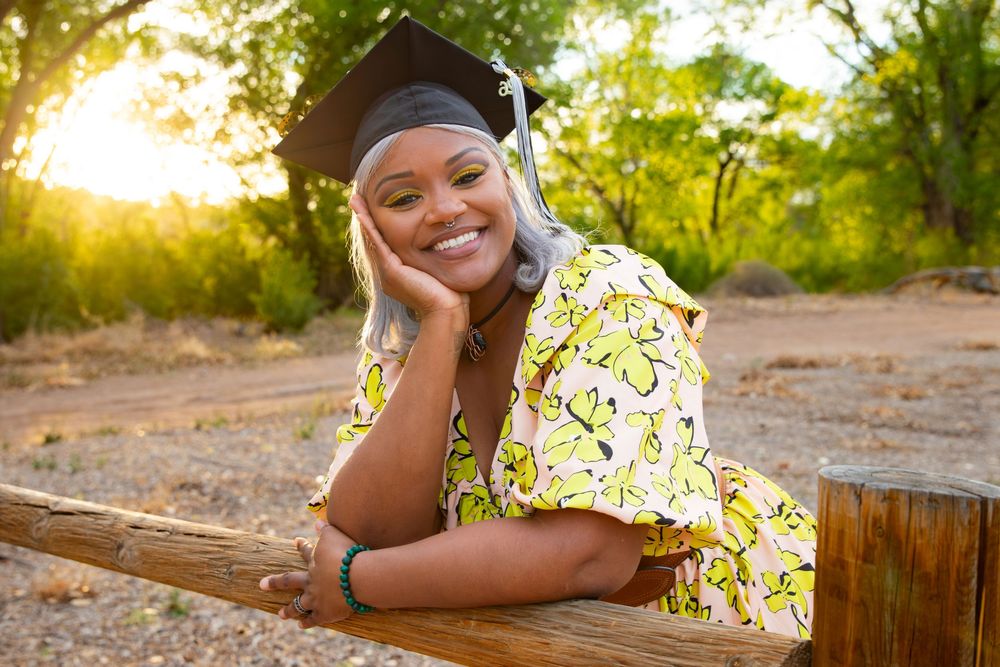 Smiling graduate in cap and floral dress posing outdoors at golden hour near wooden fence.
