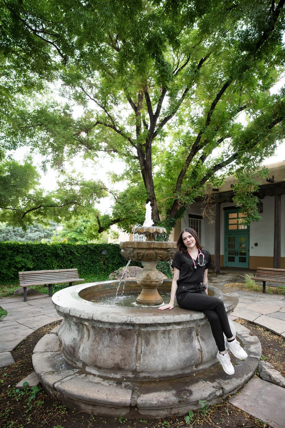 A young woman in black scrubs sits on a stone fountain in a lush garden courtyard with a large tree overhead.