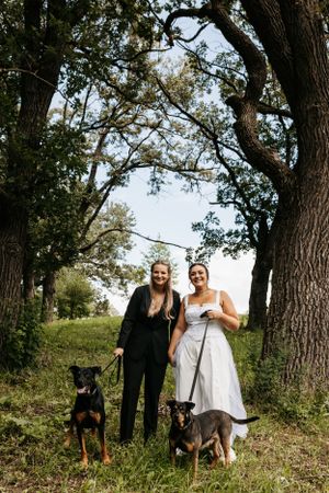 Couple poses with their two dogs under large trees in a natural outdoor setting.