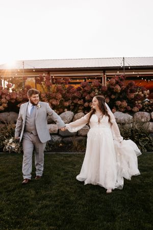 Couple holds hands while walking on grass at sunset with floral backdrop and string lights.