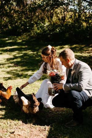 A couple in wedding attire sits on the grass feeding chickens during an outdoor photoshoot.