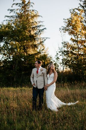 A couple stands together in a field at golden hour with tall evergreen trees in the background.