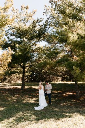 A couple shares a moment under tall pine trees in a sunlit outdoor setting.