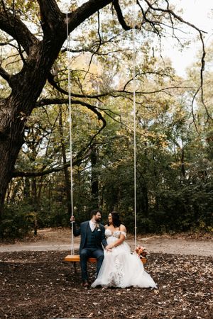 A couple enjoys a romantic moment on a wooden swing in a wooded setting.