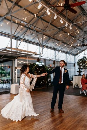 A couple sharing their first dance on a wooden floor under industrial ceiling with string lights.