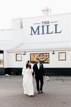 A couple in wedding attire walking outside The Mill building with their small dog on a cloudy day.