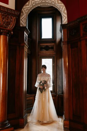 A person in a wedding gown holding a bouquet while standing in an ornate wooden alcove.