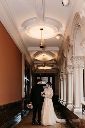 A couple embracing in an elegant hallway with ornate architectural details and pendant lighting.