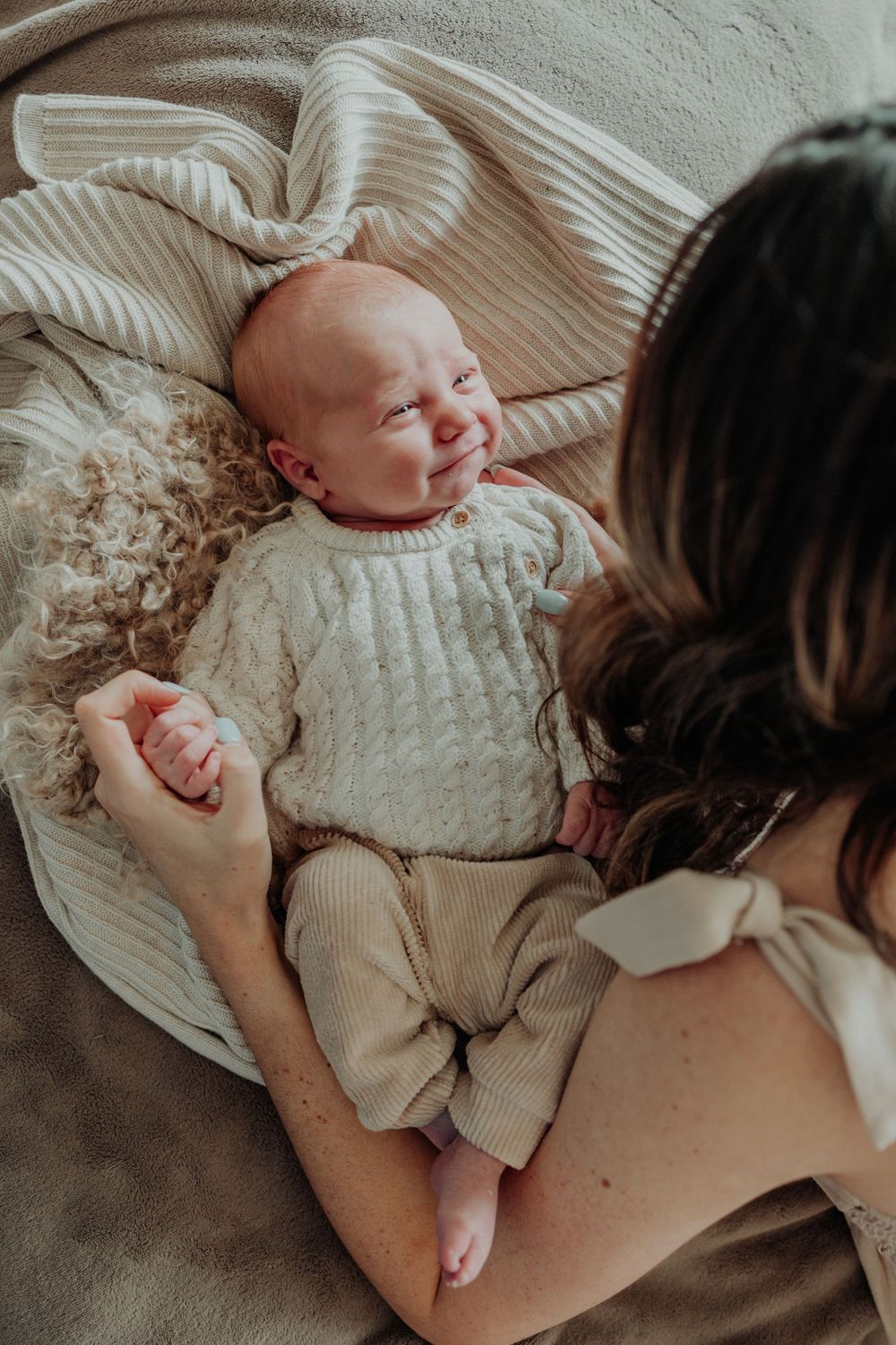 Mother looking down at smiling newborn baby dressed in cream knit outfit lying on soft blankets.