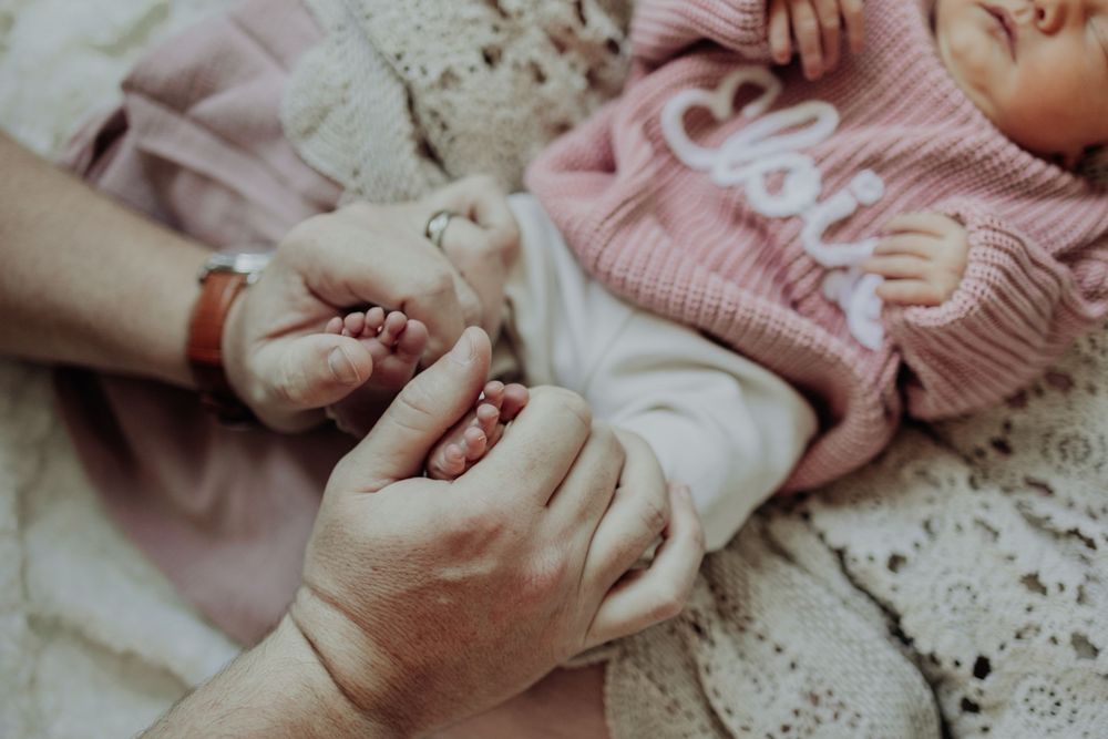 Adult hands gently holding a newborn baby's tiny feet and hand, with infant wearing a pink knit sweater.