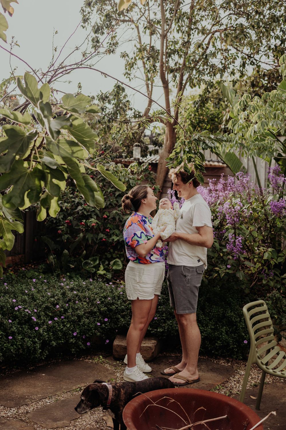 Happy couple stands together in a garden holding their newborn baby with a dog nearby.