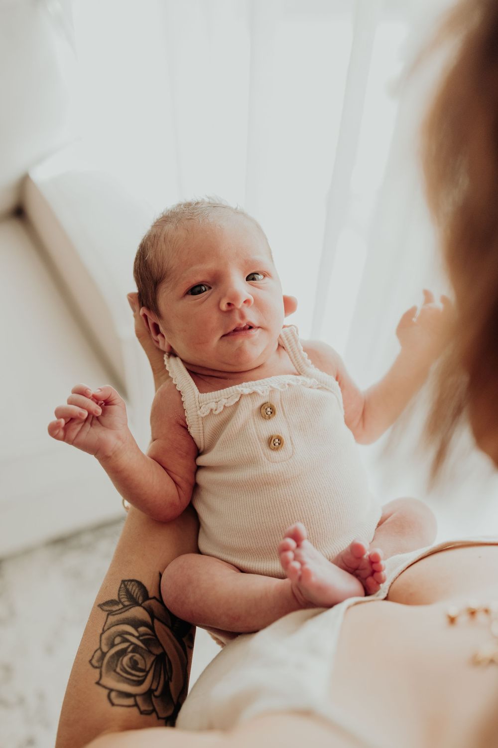 Alert newborn baby in cream romper being held up by tattooed mother in soft natural light.